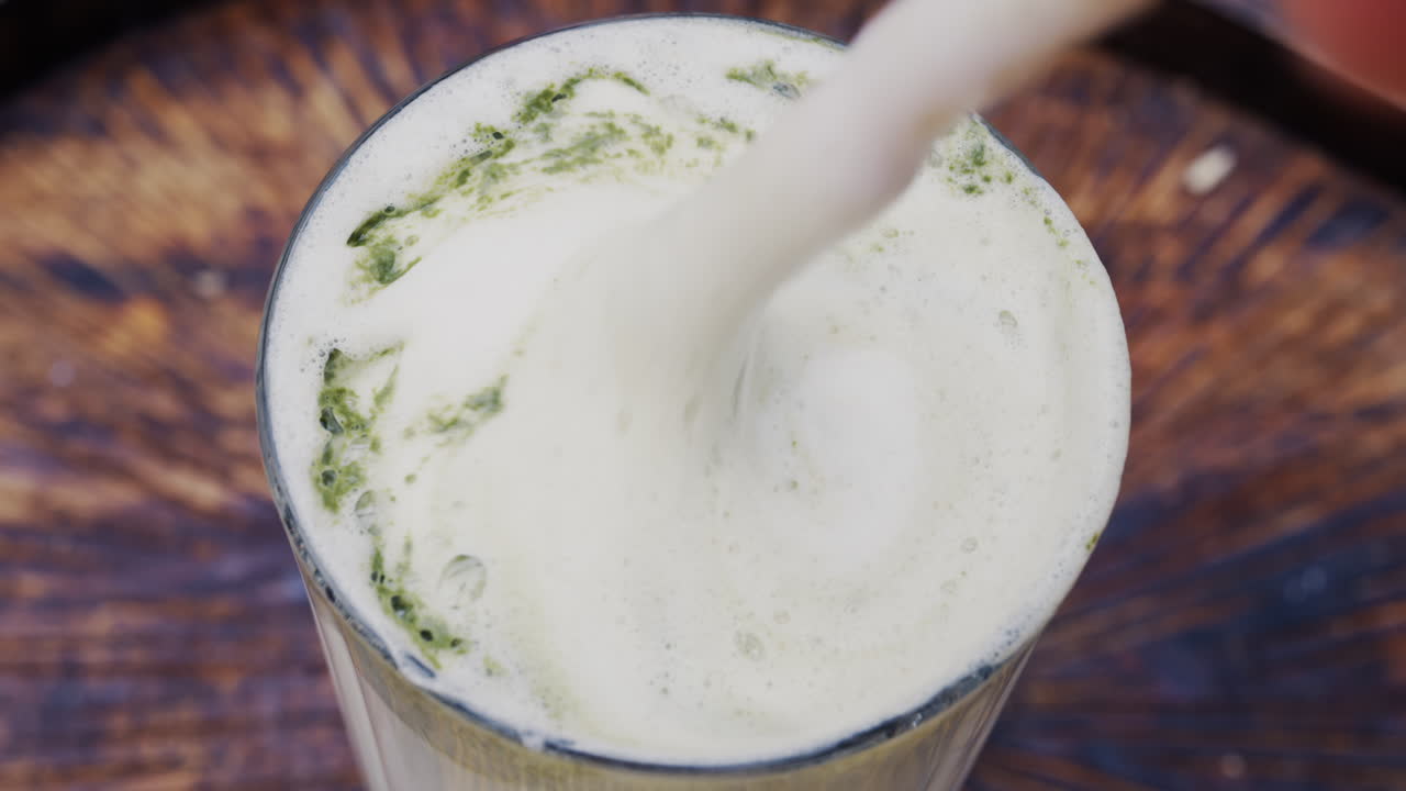Close up of a woman mixing the foam on a matcha latte with a paper straw on a wooden tray