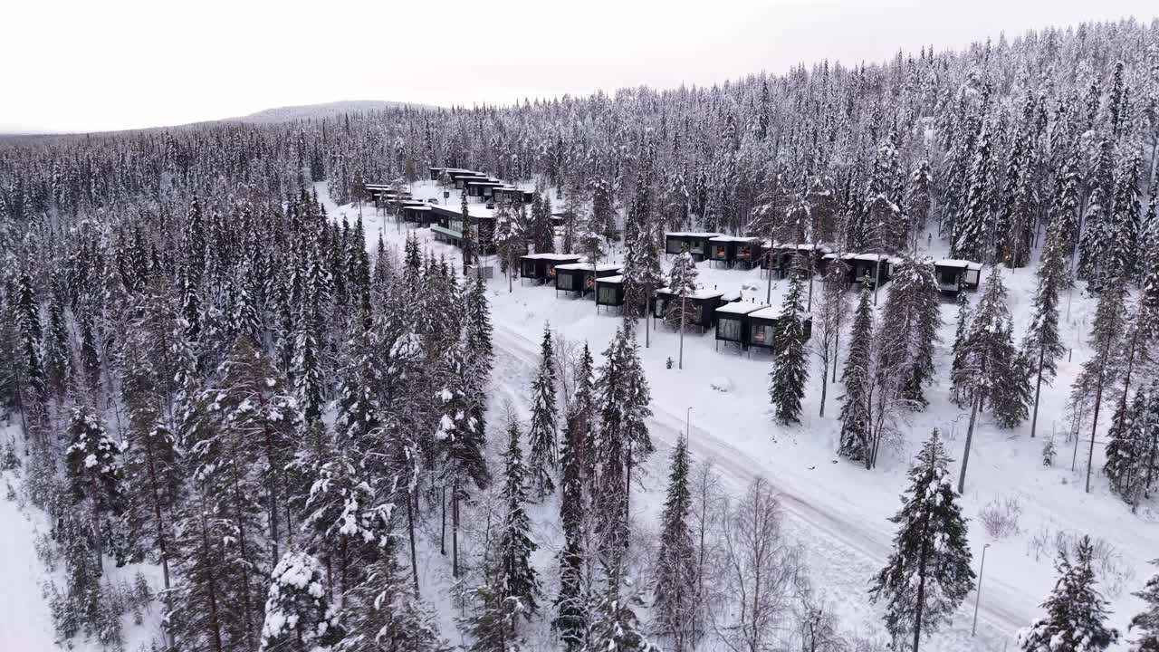 Talvinen lomakeskus, lasiset mökit mäntymetsän ympäröimänä lumipeitteen alla, Joulupukin kylä, Lappi, drone-kuva