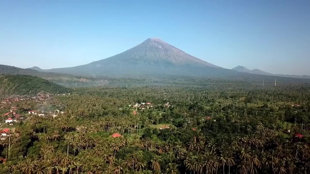 Drone video capturing Mount Agung's grandeur over dense tropical forests in Bali, Indonesia. Aerial footage provides a sweeping view of the landscape, bathed in vibrant light
