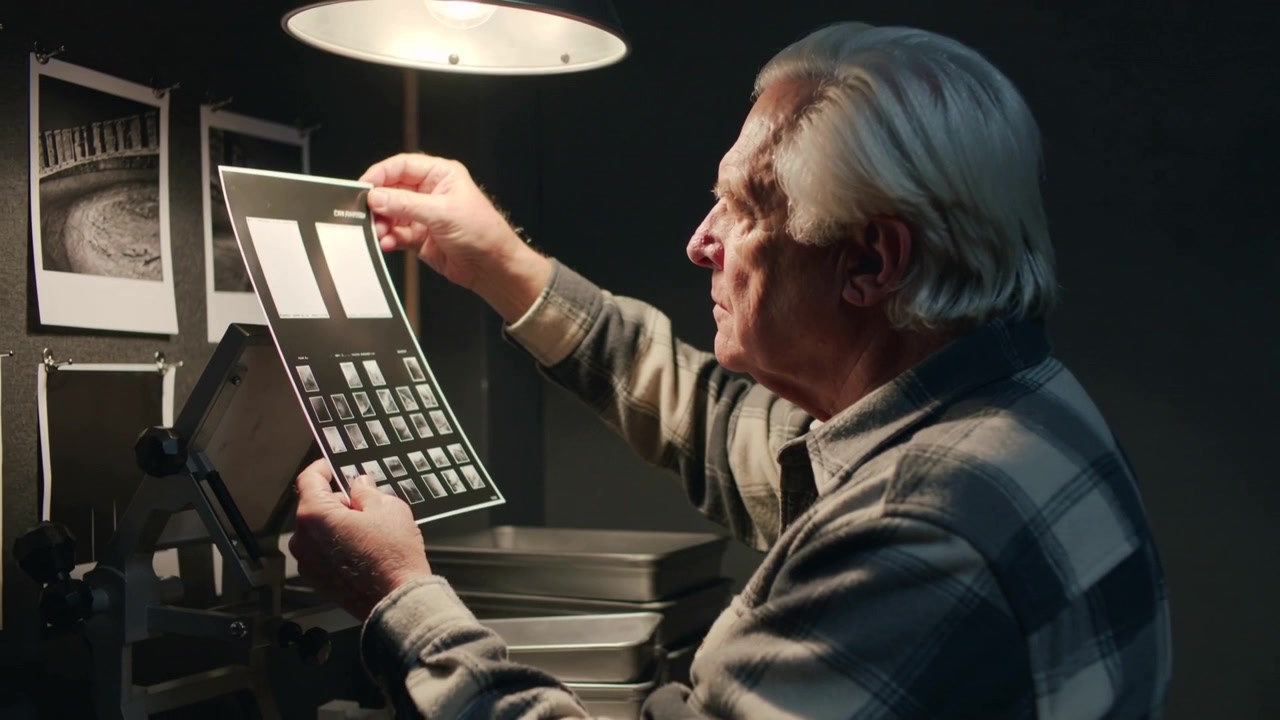 Lifting contact sheet in lab, plaid-shirt man inspecting under lamp, comparing prints, copy space