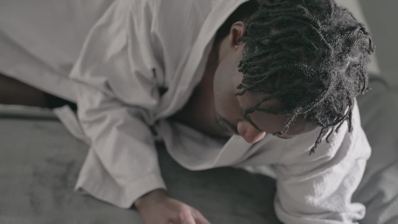 Man in white robe lying on bed, focused and relaxed, deeply immersed in reading large book, soft light streaming through window creates calm, peaceful atmosphere enhancing concentration