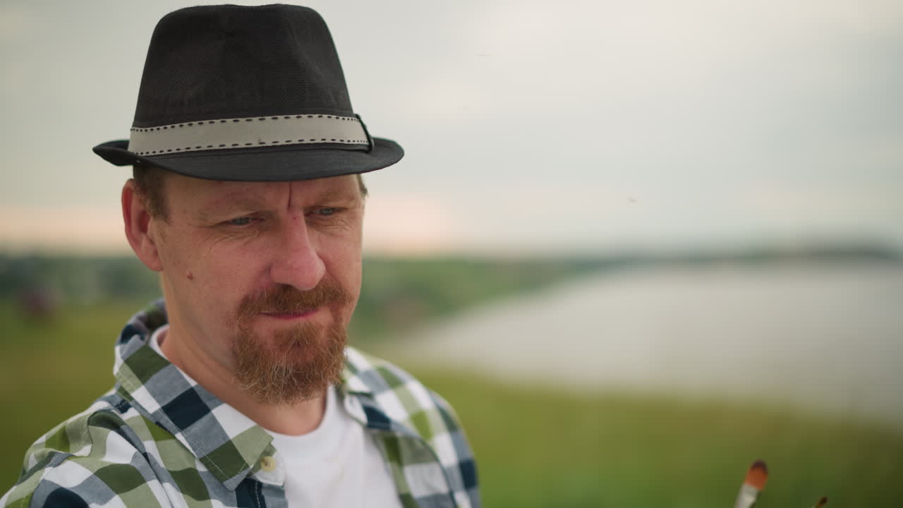 An artist is captured in a moment of deep concentration, his face serious and focused as he works on his painting in a blurry, grassy field. The background fades softly