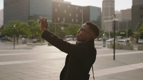 Retrato-De-Un-Estudiante-Afroamericano-De-Moda-Tomando-Fotos-De-Una-Ciudad-Urbana-Usando-Tecnología-De-Video-Para-Teléfonos-Inteligentes-Sonriendo-Disfrutando-De-Viajes-Turísticos