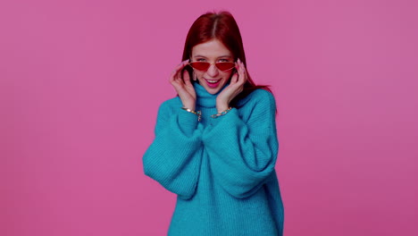 Sincere-cool-cheerful-redhead-girl-in-blue-sweater-wearing-sunglasses,-charming-smile-on-pink-wall