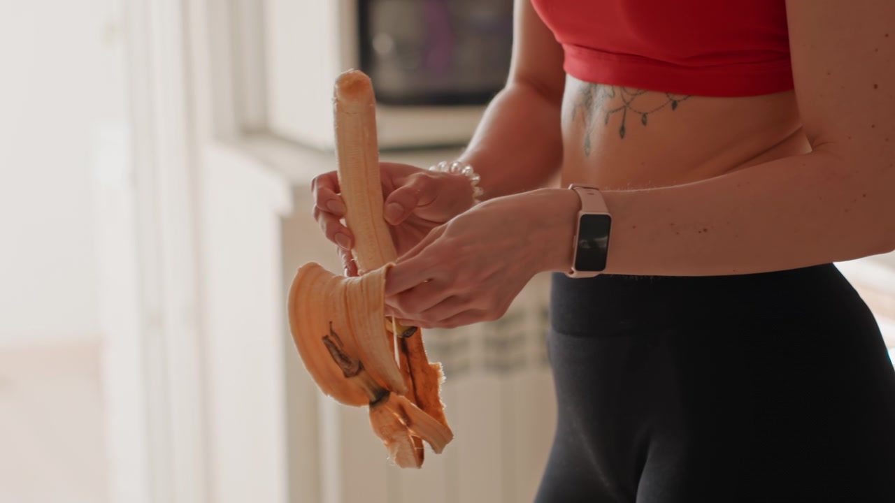 Closeup woman peeling banana in kitchen, red crop top and black leggings, smartwatch on wrist, exposed midriff, after workout snack prep, warm morning light on countertop, minimal modern kitchen