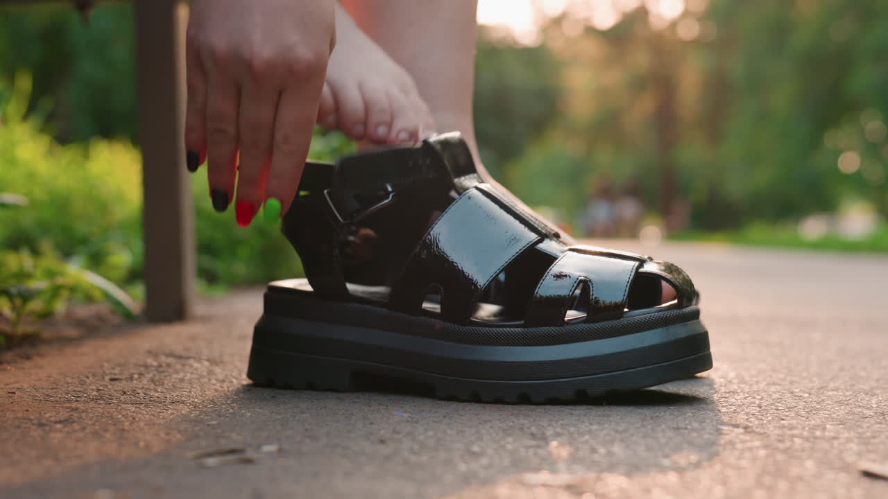 해 질 녘 산책을 준비하며 샌들을 조심스럽게 신는 우아한 여성, 신발을 고정하는 여성에게 따뜻한 햇살이 드리워져요. 여성은 저녁 산책 전에 샌들이 확실히 고정되었는지 확인합니다.