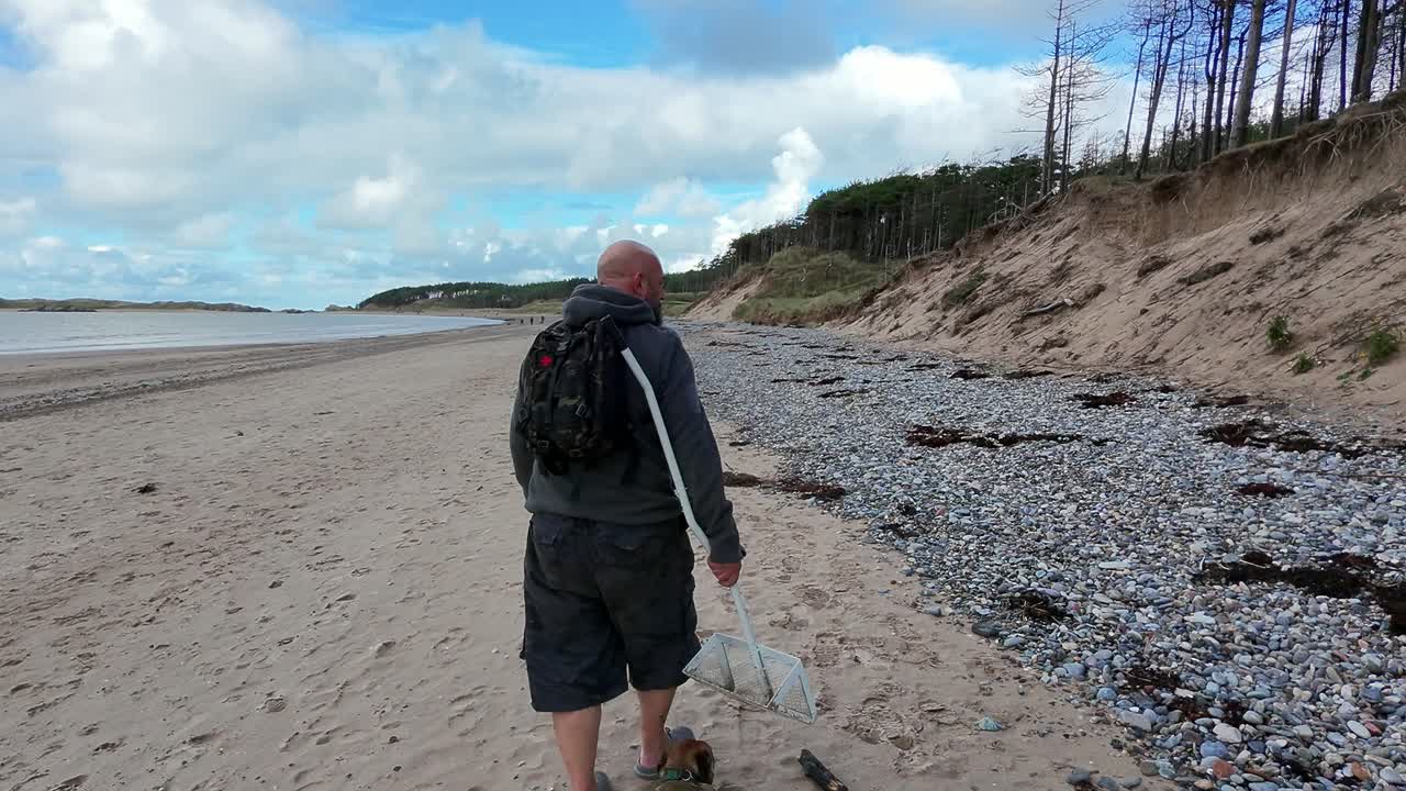 Bald backpacker male carrying sand scoop walking dog on sunny Welsh beach active lifestyle exploring