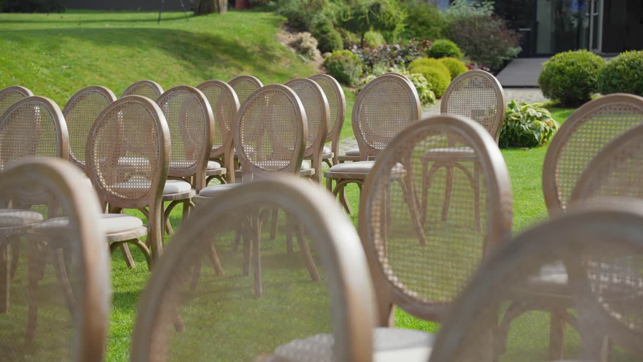 File di eleganti sedie in legno allestite all'aperto su erba verde lussureggiante, perfette per matrimoni o eventi. L'ambiente sereno del giardino e l'illuminazione naturale creano un'atmosfera pacifica.