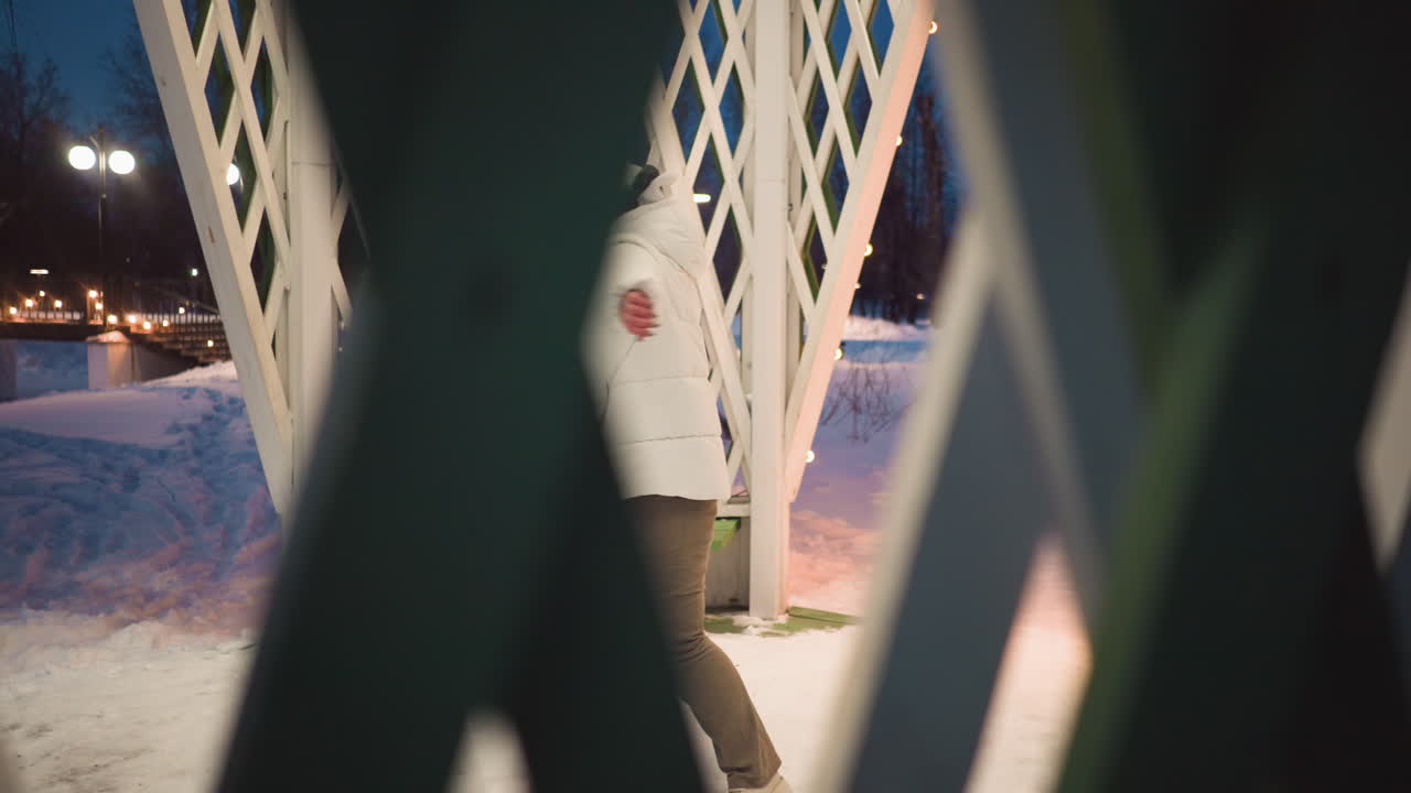 Young lady gently moves body inside lattice gazebo at dusk, wearing white puffer coat and beanie, snow covered park setting under soft lights, expressing serene dance rhythm in winter evening scene
