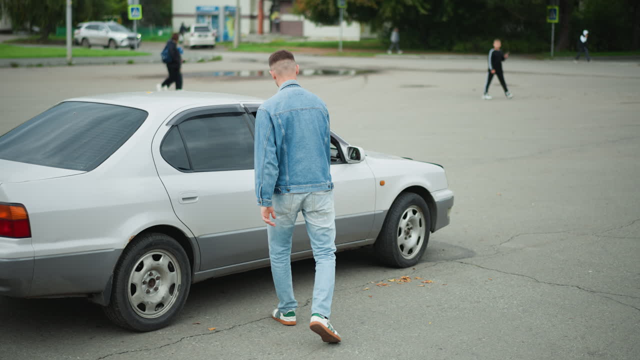 Giovane uomo bianco che si avvicina a una berlina argentata in un parcheggio vuoto, indossa una giacca di jeans, apre lo sportello del conducente e sale sul sedile, afferra il volante e prepara l'auto per la partenza, ambiente urbano determinato