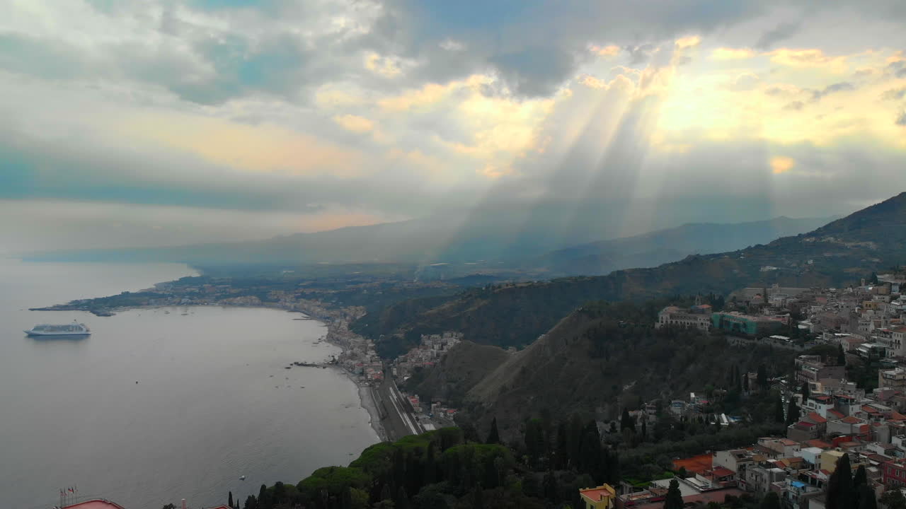 dolly aéreo en: dron volando hacia una bahía increíble con una navegación de ferry, una vista costera y un antiguo pueblo en la montaña, con un épico cielo nublado disparando rayos de luces por todas partes