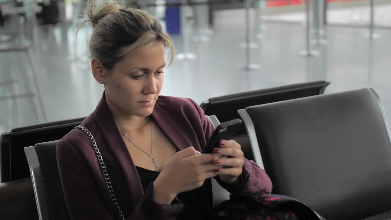 atractiva mujer caucásica escribiendo un mensaje en su teléfono inteligente mientras está sentada en la terminal del aeropuerto. ulba y feliz. el concepto de esperar el vuelo, vacaciones. cámara lenta, primer plano