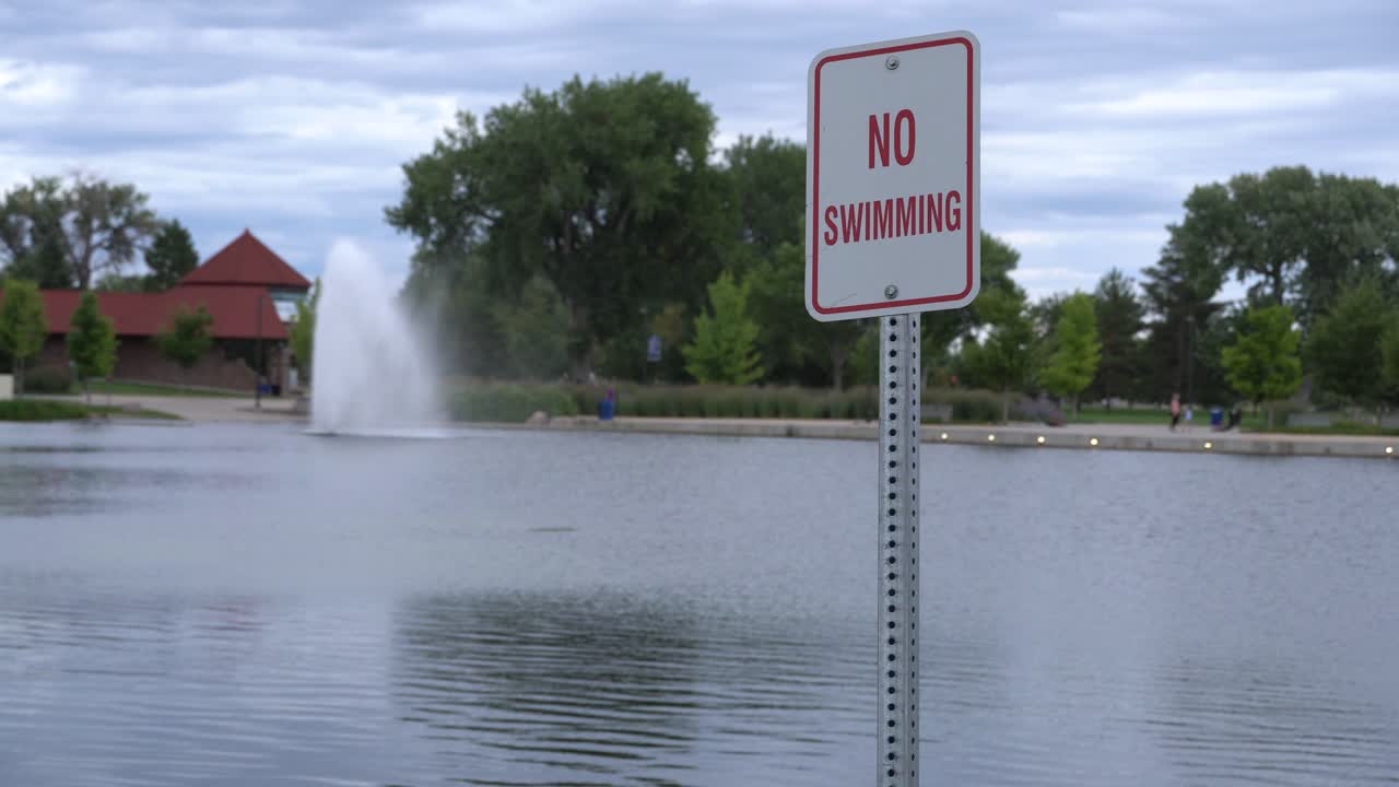 No swimming sign next to pond with fountain in background.