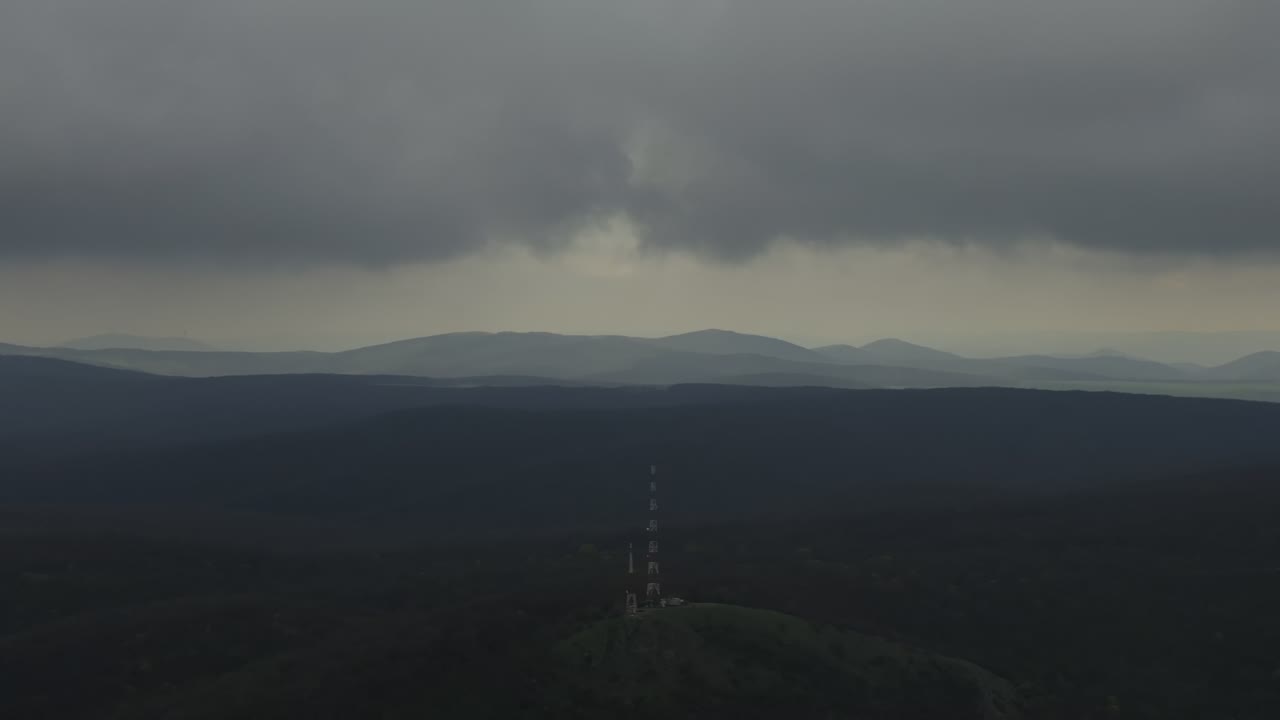 Aerial close-up pan over a layered mountain landscape under dramatic storm clouds. Soft light rays break through the heavy sky, casting subtle gradients across distant hills