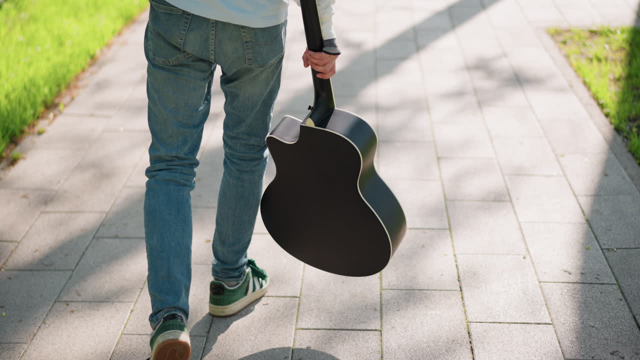 gitarist, güneşli park kaldırımında akustik gitarıyla yürürken, kot pantolon ve spor ayakkabı giymiş, rahat adımlarla uzun gölgeler oluşturuyor, enstrümanı sapından tutmuş, konser yolunda, düşünceli bir ruh hali
