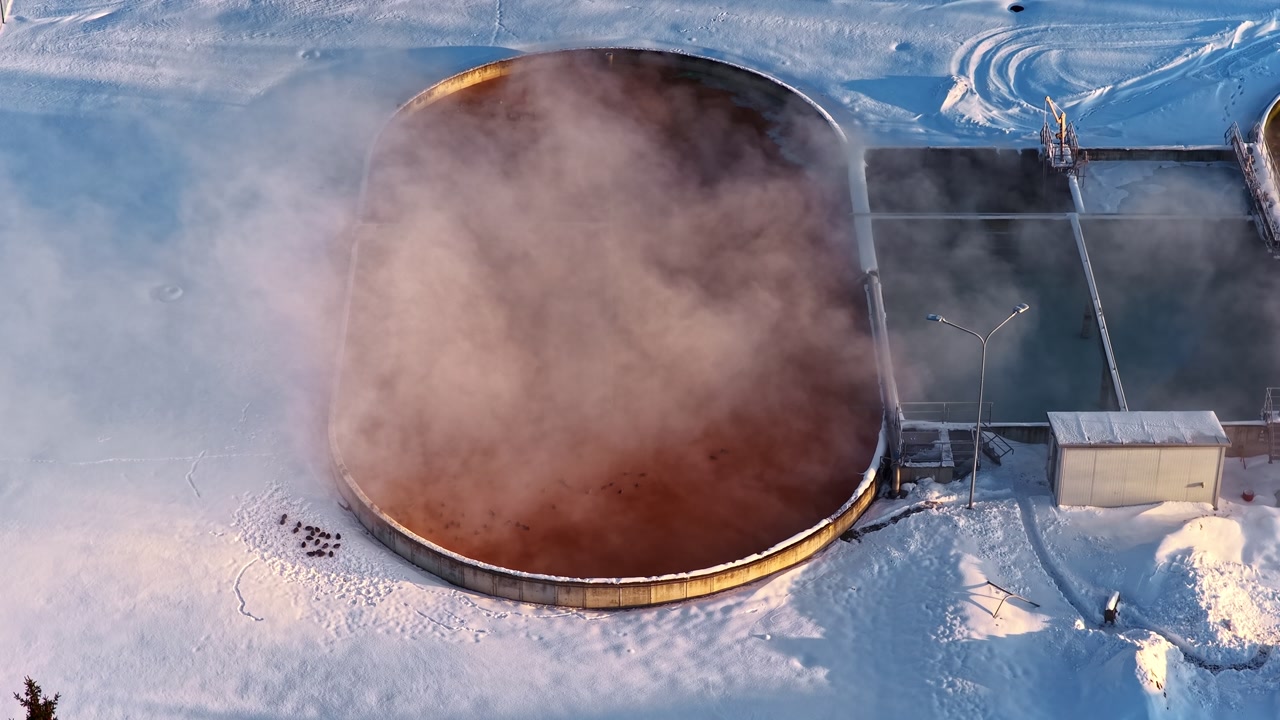 Aerial drone view of large circular industrial wastewater treatment tank with steam rising over snow-covered facility grounds in winter Latvia