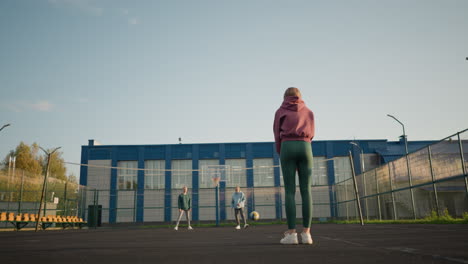 two women on other side of volleyball court, lady in cyan rolls ball back to lady in maroon hoodie and green leggings. building and outdoor court visible in background