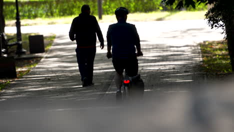 Backlit-view-of-a-cyclist-and-pedestrian-on-a-sun-dappled-park-path,-conveying-a-serene,-urban-outdoor-activity