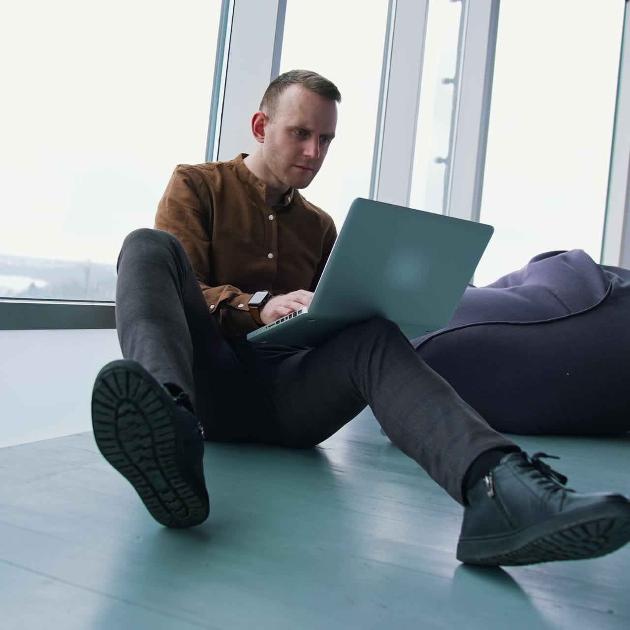 Confident man working on a laptop. Young businessman sitting on a floor and typing on a wireless computer near the window at daytime. Serious freelancer works indoors