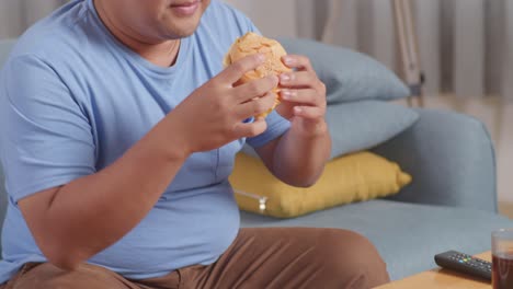 close up of a fat asian man smelling to hamburger in his hands then smiling while eating fast food on a sofa in the living room at home