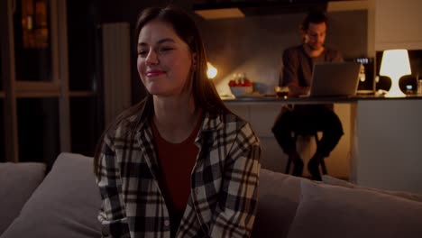 una ragazza bruna calma e interessata con una camicia a scacchi si siede su un divano grigio e guarda la tv passando i canali usando il telecomando mentre il suo ragazzo lavora su un portatile in cucina in un appartamento in studio la sera