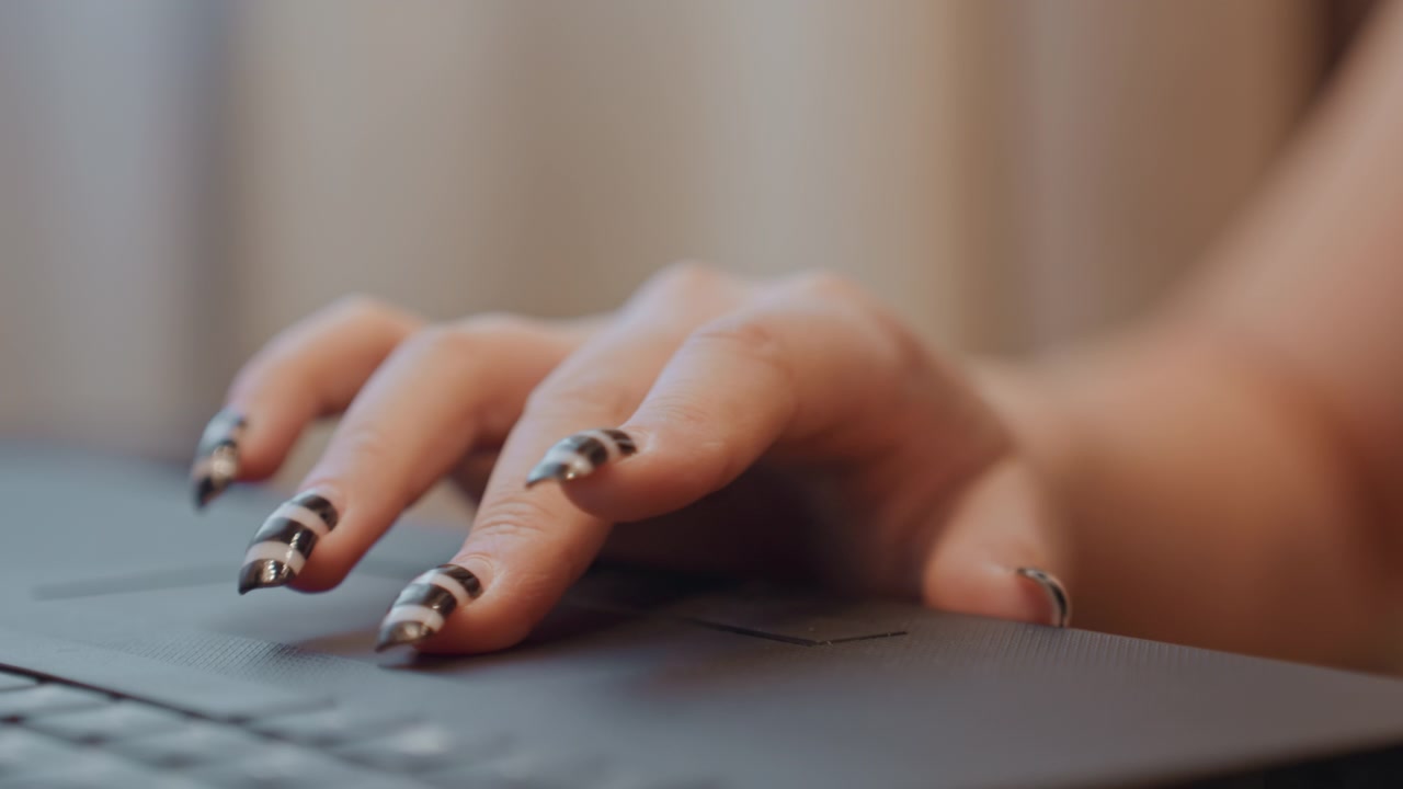 Primo piano di mani che digitano su un laptop con una manicure a strisce audaci, unghie lucide bianche e nere, influencer che modifica contenuti social in uno studio accogliente, battiture lente e deliberate e umore concentrato