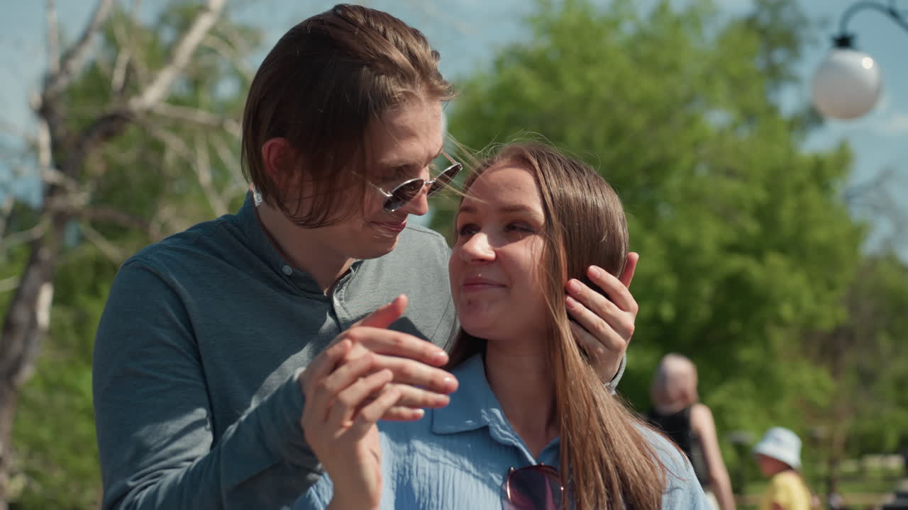 선글라스를 쓴 남자가 야외에서 눈을 가리고 부드럽게 키스하며 백인 여자친구를 놀라게 하는 장면, 가로등과 걸어 다니는 사람들로 붐비는 공원, 푸른 덮개, 편안한 포옹, 솔직한 미소와 따뜻함