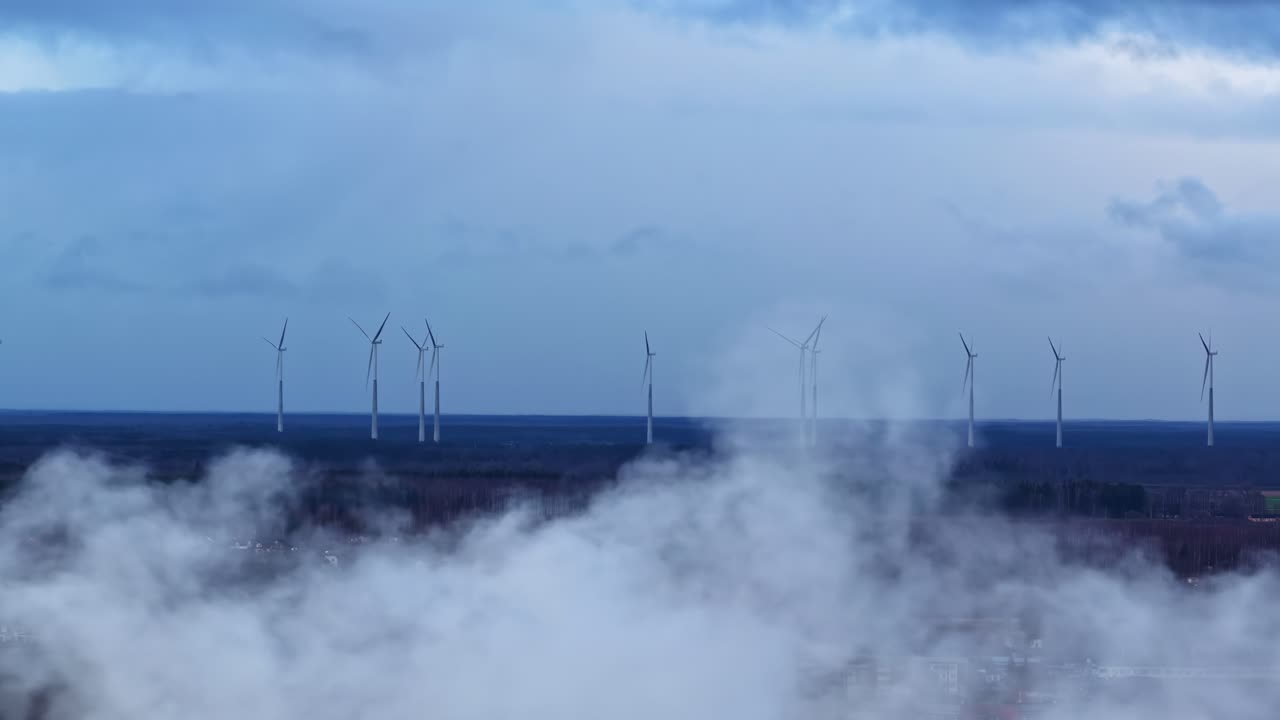 Letecký záběr větrných turbín tyčících se nad zamlženou průmyslovou krajinou s městskými budovami a vzdálenou větrnou farmou pod zataženou modrou oblohou