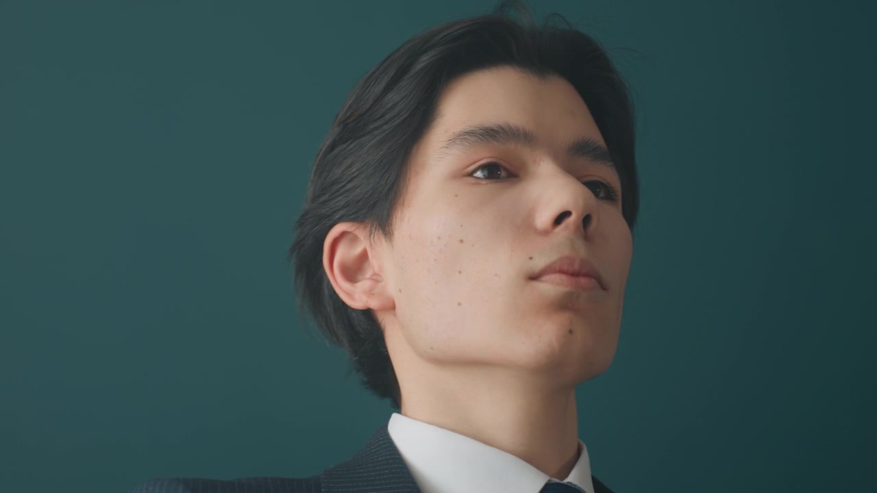 スーツを着た若いアジア人男性が髪を整え、ネクタイを直している。ティール色の背景、柔らかなシネマティックな照明、落ち着いた表情のエディトリアルポートレートシリーズ。プレゼンテーションとリーダーシップのために準備するモデル。