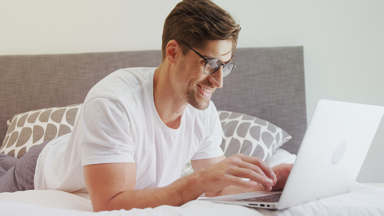 Smiling man wearing spectacles lying on bed using his laptop 4K 4k