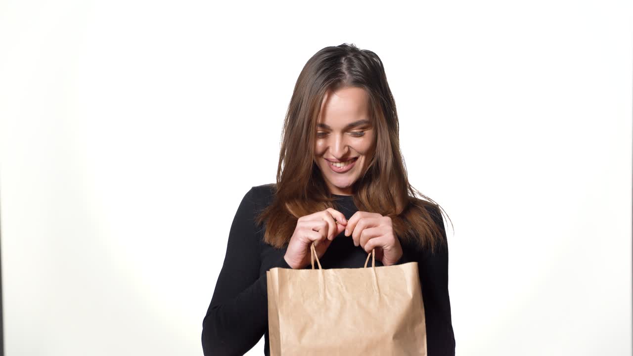 A playful brown-haired woman with long hair holds a craft package with a gift in her hands and looks inside it on a white background. Shopping.