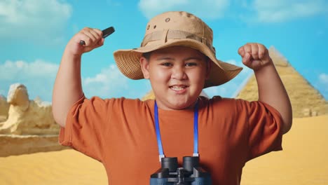 asian boy with a hat and binoculars using the magnifying glass then flexing his bicep while traveling in giza pyramid. boy researcher examines something, travel tourism adventure, close up
