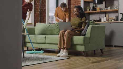 afroamerican girl using laptop sitting on sofa while roommates cleaning the house