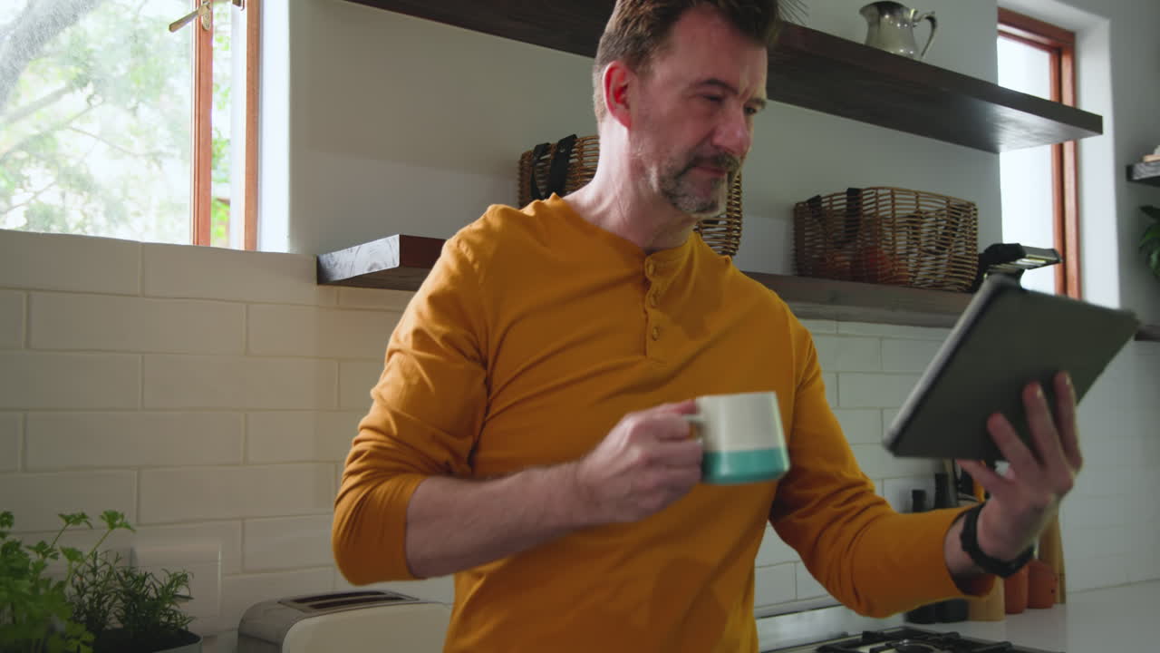 Uomo anziano in cucina che si gode un caffè mentre legge sul tablet, sentendosi rilassato, a casa.