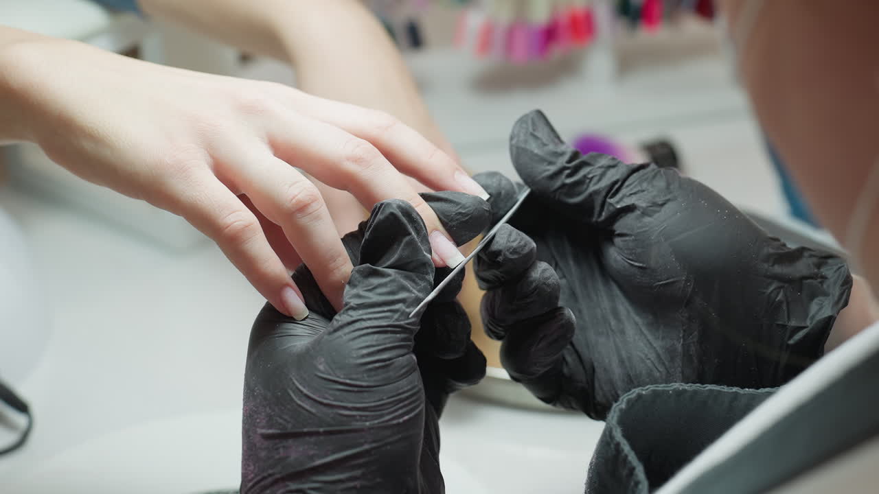Close up image of nail technician wearing black gloves filing customer's fingernail with precision during manicure session at salon, with soft focus background showing tools and colorful nail samples