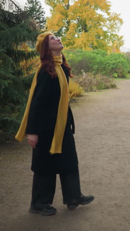 joyful lady with long red hair, wearing a yellow beret, scarf, and sunglasses, happily turning around as she walks in a garden with autumn foliage, embodying the essence of fall