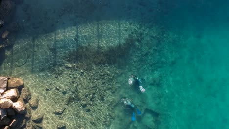 a high view looking down at 2 scuba divers swimming in clear water next to a rocky outcrop