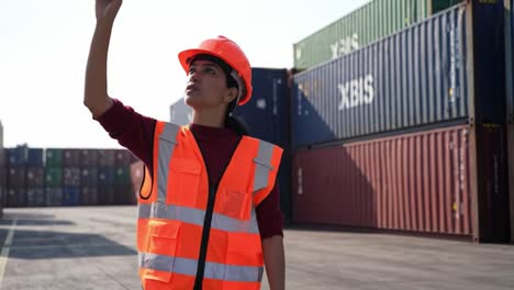 a warehouse worker assesses the area in an industrial yard, surrounded by shipping containers, demonstrating strong safety protocols with a bright vest and helmet while engaged in her duties.
