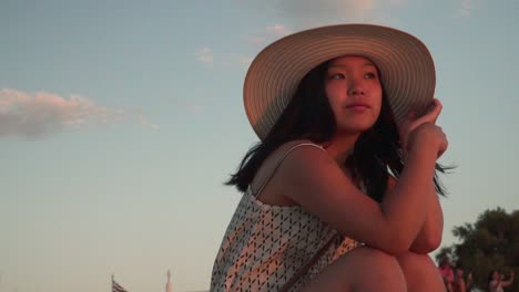 Girl-with-summer-hat-sitting,-looking-into-distance-with-backdrop-of-small-greek-flag-and-clouds-SLOW-MOTION