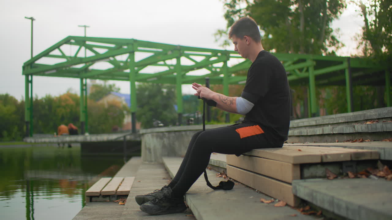 Valkoihoinen miespuolinen videobloggaaja säätää kameran jalustaa penkillä lammen lähellä valmistautuessaan kuvaamaan käsivaralta vihreän pergolan alla, luonnonvalo, lähikuva käsistä, hihnasta ja varusteista, huolellinen asettelu, kaupunkimainen ranta