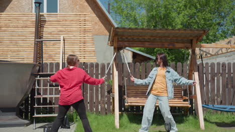 two women dressed casually engage in playful sword fight in sunny backyard near wooden swing and fenced area, striking lighthearted poses while enjoying outdoor fun on paved walkway