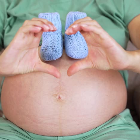 big belly of pregnant woman where she puts tiny knitted socks. future mommy shows heart sign with her hands. beautiful little baby socks on the belly closeup