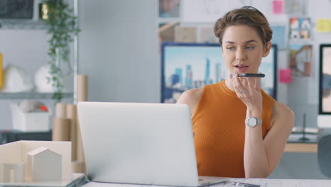 Female-Architect-In-Office-Working-At-Desk-On-Laptop-Taking-Speakerphone-Call-On-Mobile-Phone