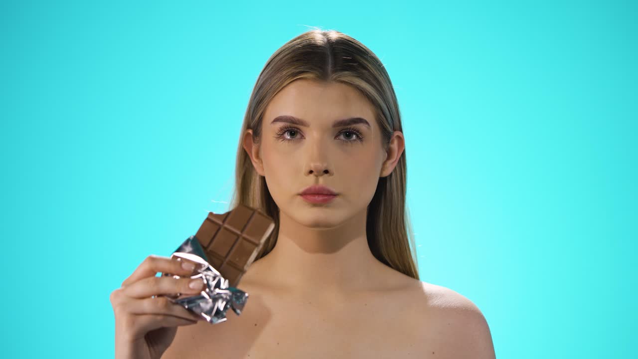 toma media de una mujer joven y atractiva mientras disfruta de un bocado de la barra de chocolate vegana y después se acaricia los labios con el dedo frente a un fondo turquesa en cámara lenta