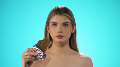 medium shot of a young attractive woman while she enjoys a bite of the vegan chocolate bar and afterwards strokes her lips with her finger in front of turquoise background in slow motion