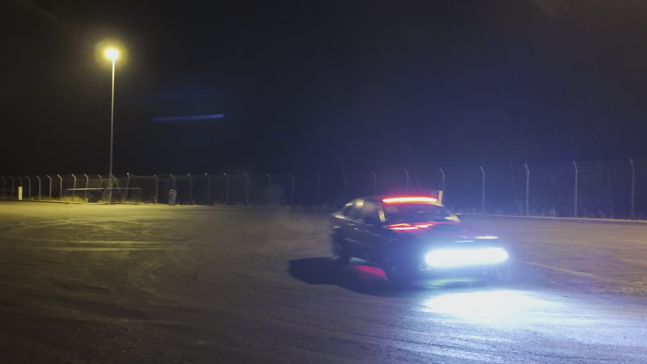 A fast sports car performing a drift in an open lot illuminated by bright streetlights on a rainy night