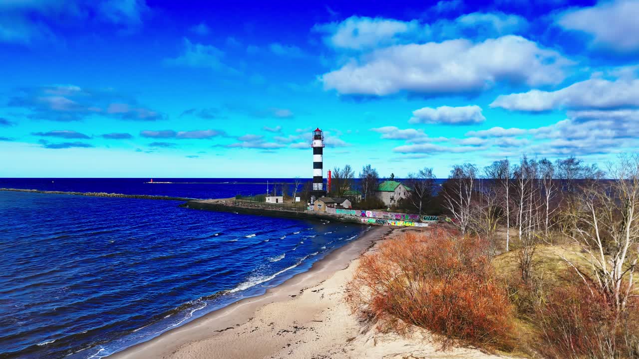 A bold striped lighthouse stands near deep blue waves and a pale sandy beach, framed by leafless trees and colorful autumn shrubs under a bright sky.