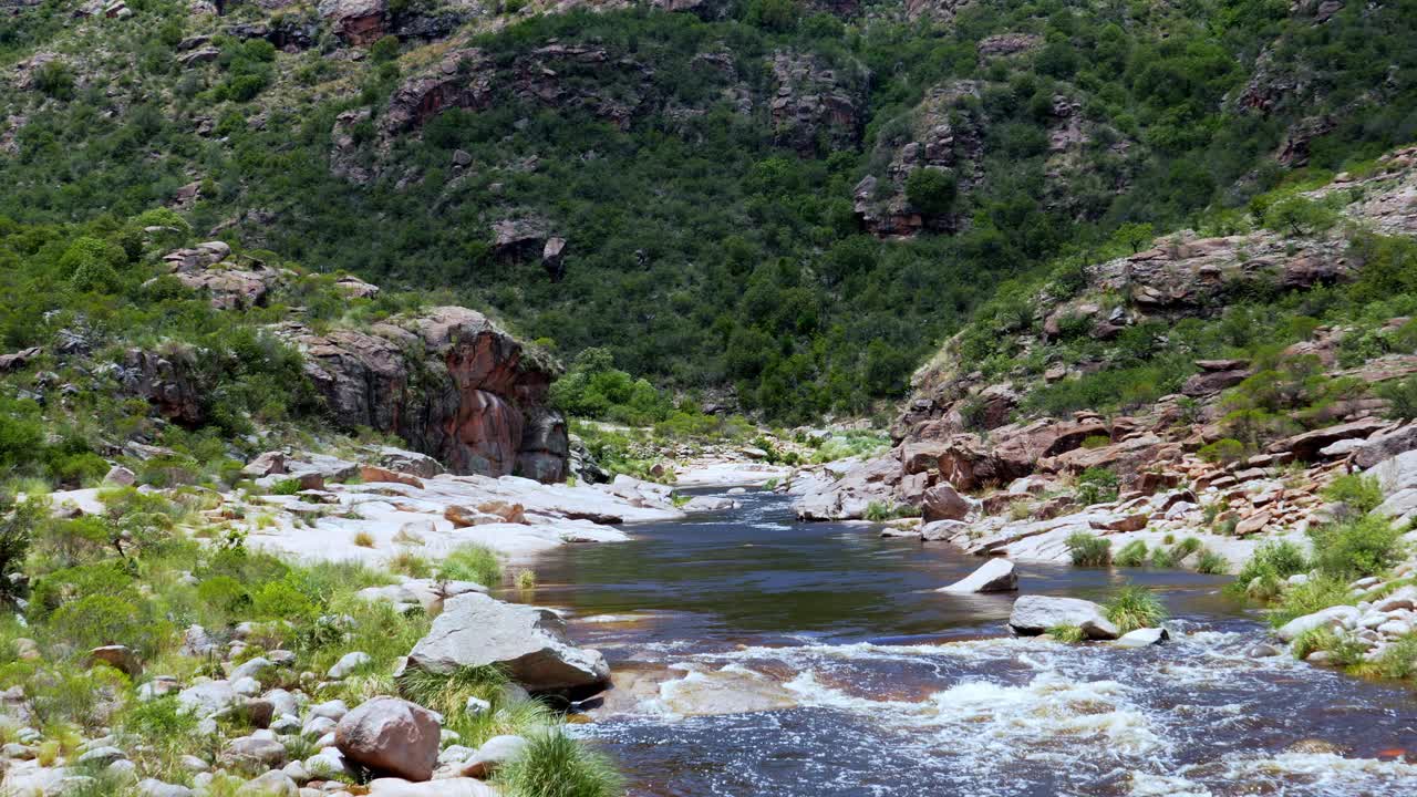 Static wide shot of Mina Clavero river flowing through a rocky canyon in the Córdoba region. Water moves between stones and vegetation, with steep green hills surrounding the riverbed. Slow-motion