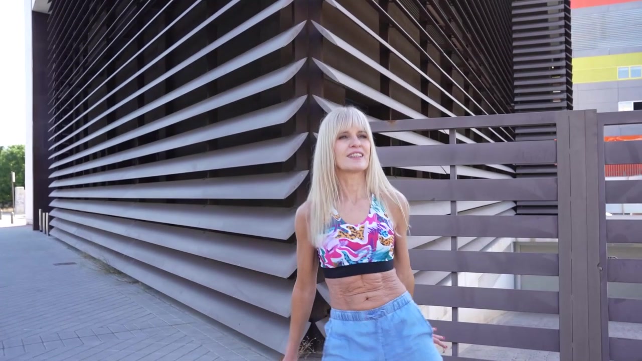 Donna bionda con top colorato e jeans che cammina vicino a un moderno edificio architettonico.