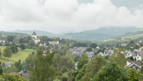 Zeitraffer-Eines-Tals-Im-Sauerland,-Deutschland,-Mit-Wolken,-Die-Am-Dorf-Grafschaft-Vorbeiziehen,-Mit-Freiem-Sichtblick-Auf-Das-Kloster-Und-Die-Typischen-Fachwerkhäuser-Vor-Einer-Abfallenden-Berglandschaft